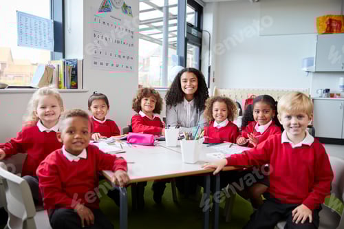 Visualização: Retrato de professora infantil e crianças sentadas à mesa em uma sala de aula olhando para a câmera sorrindo