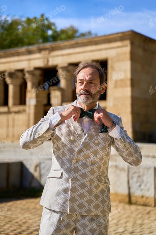Preview: Smiling man adjusting bow tie near ancient stone temple outdoors