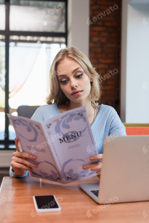 Preview: young blonde woman reading menu near laptop and smartphone with blank screen