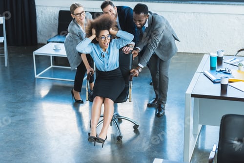 Preview: multicultural businesspeople having fun and racing colleague on chair in office