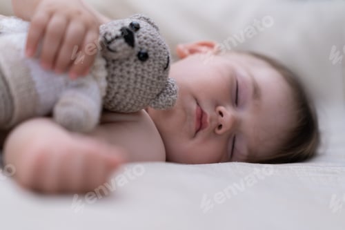 Preview: Adorable chubby baby hugging knitted bear toy with hand lying on soft bed in nursery