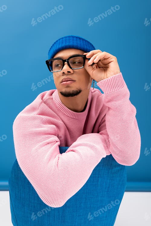 Preview: Handsome young man in stylish pink sweater and blue hat poses confidently against bright background
