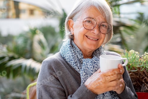 Preview: Portrait of happy senior woman smiling drinking hot coffee or tea standing outdoor on the balcony