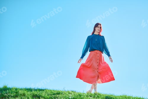 Preview: Teenage girl in a blue blouse and orange skirt standing on grass against a clear blue sky.