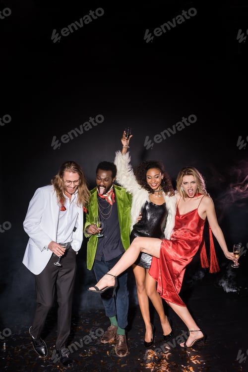 Preview: happy multicultural friends standing with champagne glasses near shiny confetti on black with smoke