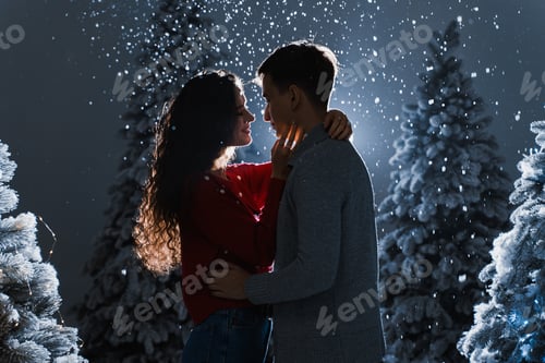 Preview: Snowy kisses on dark blue background. Young couple hugs near Christmas trees before New Year.
