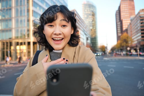 Preview: Portrait of asian girl looks surprised at smartphone screen, video chats and hears amazing news