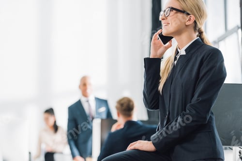 Preview: smiling young businesswoman in eyeglasses talking by smartphone and looking away in open space