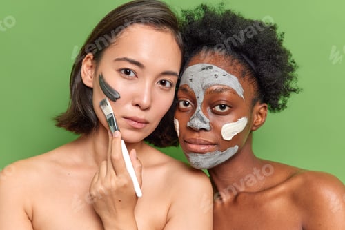 Preview: Portrait of diverse young women look seriously at camera apply clay mask use cosmetic brush stand to