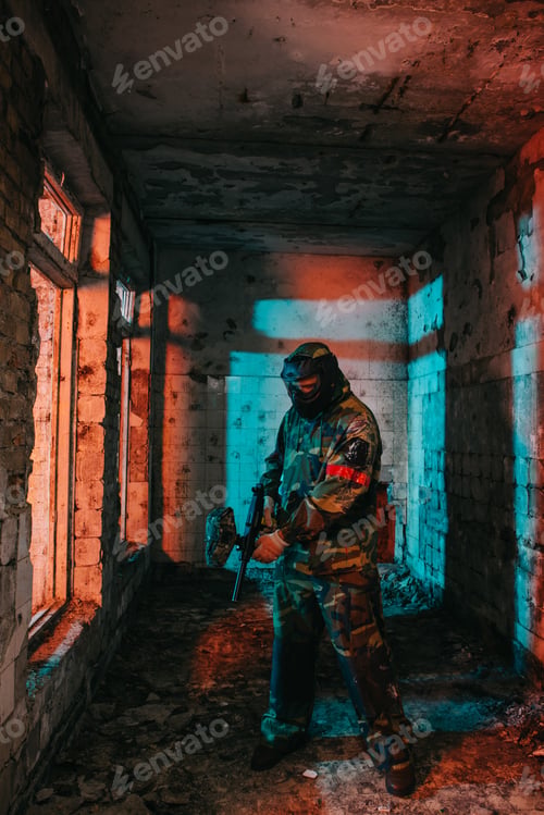 Preview: male paintball player in goggle mask and camouflage loading paintball gun in abandoned building