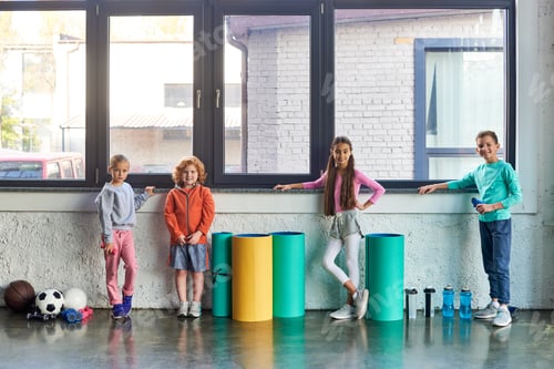 Preview: four preadolescent cute boys and girls in sportswear posing by window in gym, child sport