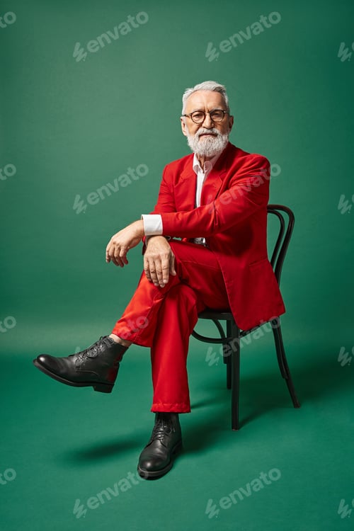 Preview: classy good looking Santa in red suit sitting on chair on dark green backdrop, winter concept