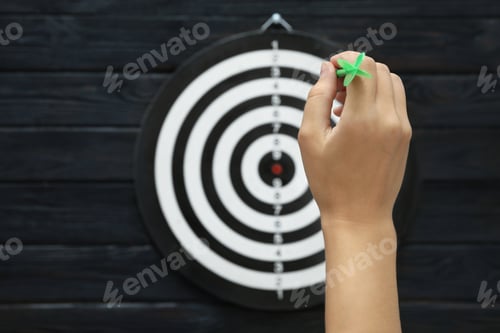 Preview: Woman throwing green arrow at dart board on dark wooden background, closeup