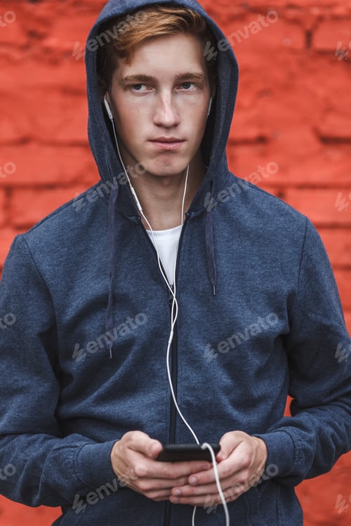Preview: Young man with cell phone and earphones at red brick wall