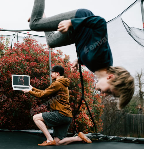 Preview: Dad holding the laptop during video chat while a young child jumps on trampoline in autumn / fall