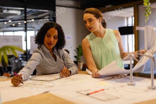 Preview: Diverse female architects in discussion using blueprints in casual office meeting