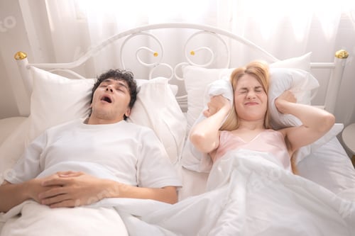 Preview: A young woman covering her ears with a pillow while her partner snores loudly
