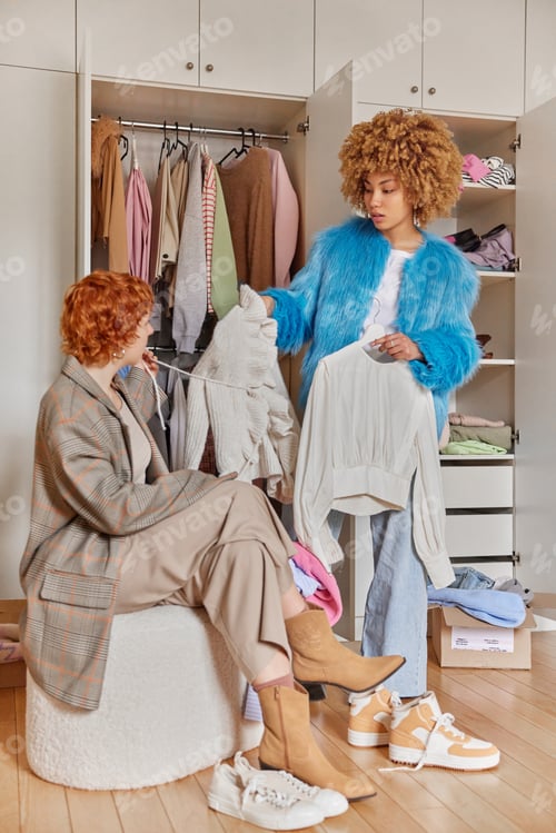 Preview: Curly haired woman holds two clothes items on hangers suggests friend what to wear stand near