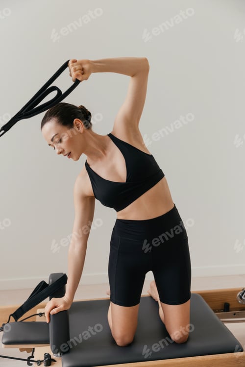 Preview: Slim brunette woman in black tight sportswear working out on Pilates reformer machine