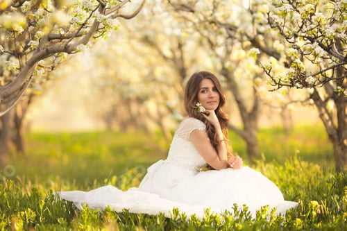 Preview: Beautiful girl in a wedding white dress sitting in a blooming garden and enjoying flowers aroma