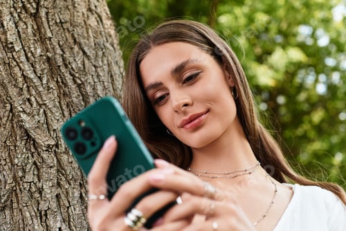 Preview: Young beautiful woman enjoying a sunny day while using her smartphone in a lush outdoor setting