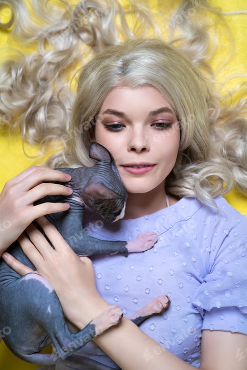 Preview: Portrait of young woman lying down on yellow background, gently clutching Sphinx kitten to her chest
