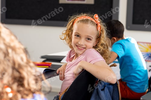 Preview: In school, young Caucasian girl with curly hair is smiling, sitting at a desk in a classroom