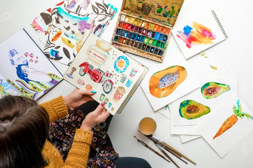 Preview: top view of woman holding album surrounded by color drawings and drawing utensils