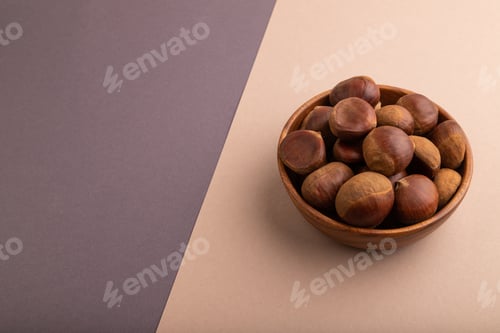 Preview: wooden bowl with raw edible Сhestnuts on brown and gray paper pastel, side view, copy space