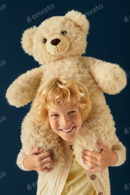 Preview: Blonde boy with wavy hair joyfully hugs a large teddy bear in studio setting