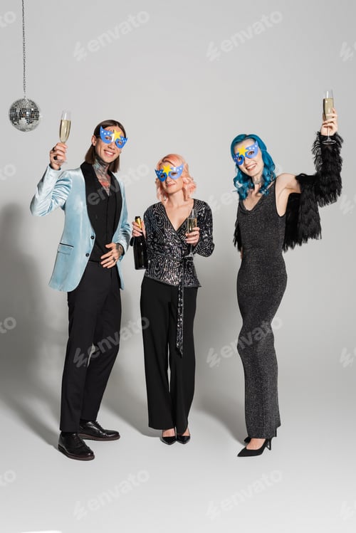 Preview: full length of stylish queer friends toasting with champagne and looking at camera on grey