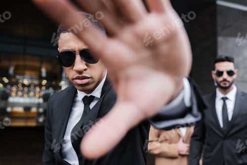 Preview: bi-racial guard in sunglasses showing stop gesture near businesswoman and african american security