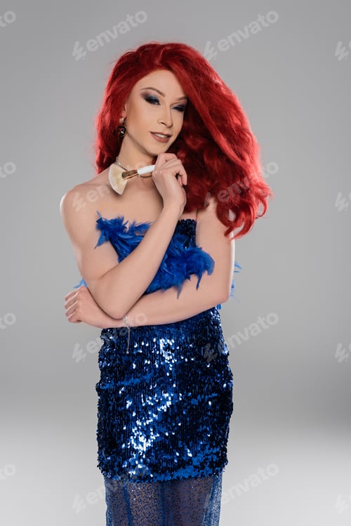 Preview: Smiling drag queen in dress holding cosmetic brush near shoulder isolated on grey