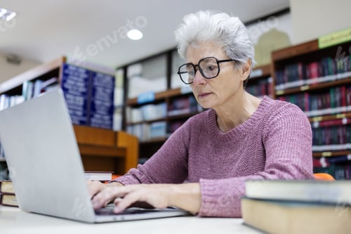 Preview: An elderly woman with gray hair and glasses works on a laptop in a library.