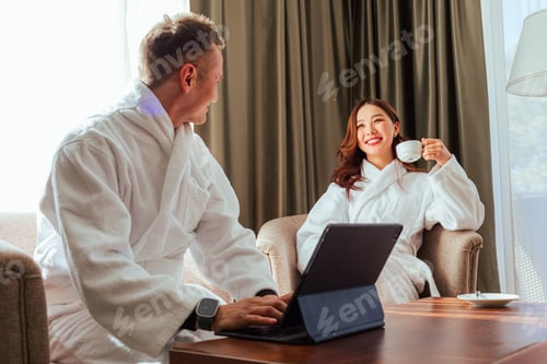 Preview: Asian couple in white robes sitting on a chair during honeymoon in hotel room