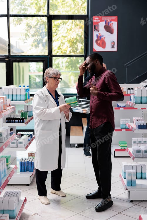 Preview: Eldely pharmacist offering pills packages to sick customer with paintful headache in pharmacy