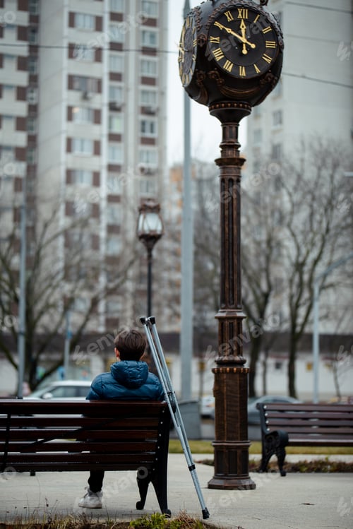 Preview: A physically disabled young man is sitting on a bench on the street near a large clock with crutches