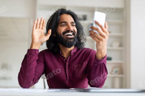 Preview: Smiling Indian freelancer man waving hand while having video call on smartphone