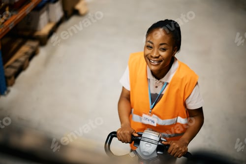 Preview: Above view of happy black female worker using pallet jack at distribution warehouse.
