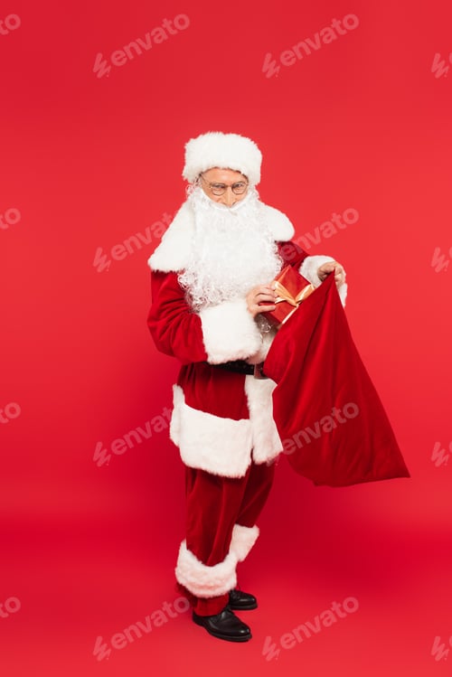 Preview: Santa claus in costume holding sack and present on red background