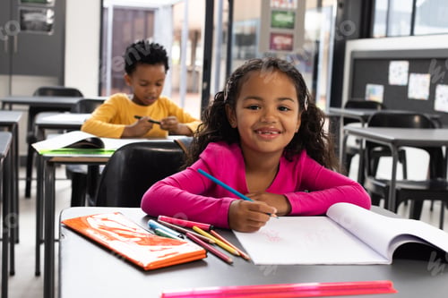 Preview: A biracial girl with curly hair smiles while drawing in a school classroom, wearing a pink top