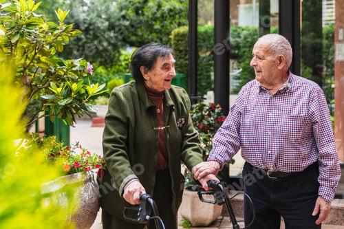 Preview: Elderly couple walking with rollators in outdoor nursing home garden