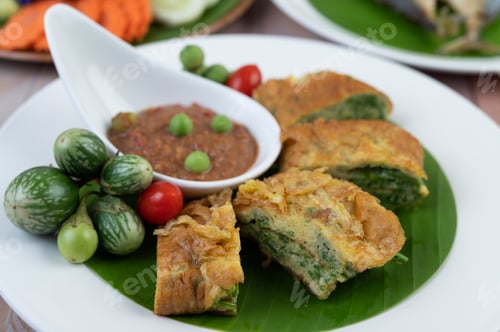 Preview: Shrimp paste, along with omelette, Cha-om and eggplant, placed in a white plate on a wooden floor.