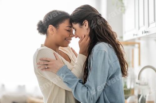 Preview: curly multiracial woman hugging cheerful girlfriend with closed eyes at home
