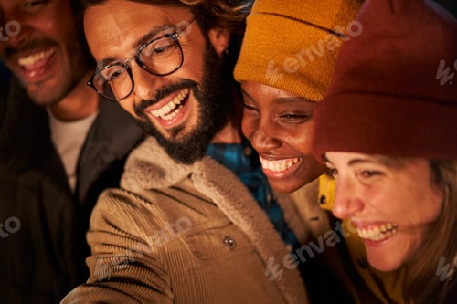 Preview: Young adults together smiling and taking selfie. Multiracial people enjoying night party in winter.