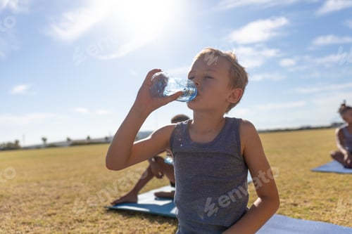 Preview: Caucasian elementary schoolboy drinking water while standing on field against bright sky