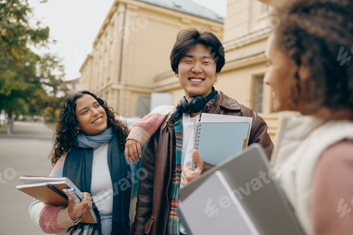 Preview: Happy students talking each other standing near university campus and holding books and laptop