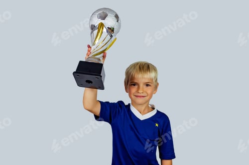Preview: Young soccer player in blue jersey holds winners cup after the goal, isolated at light background
