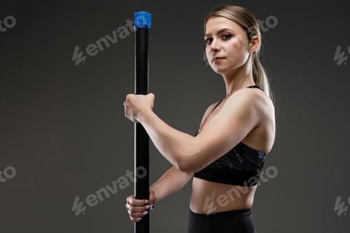 Preview: strong girl in a sports top holds a gymnastic stick and stands half-sided against a dark background