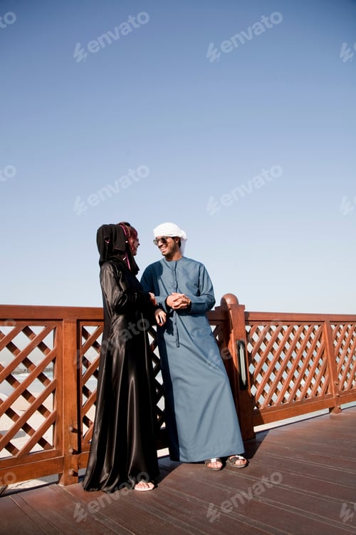 Preview: Two people in traditional Emirati attire sharing a joyous moment on a sunny boardwalk.